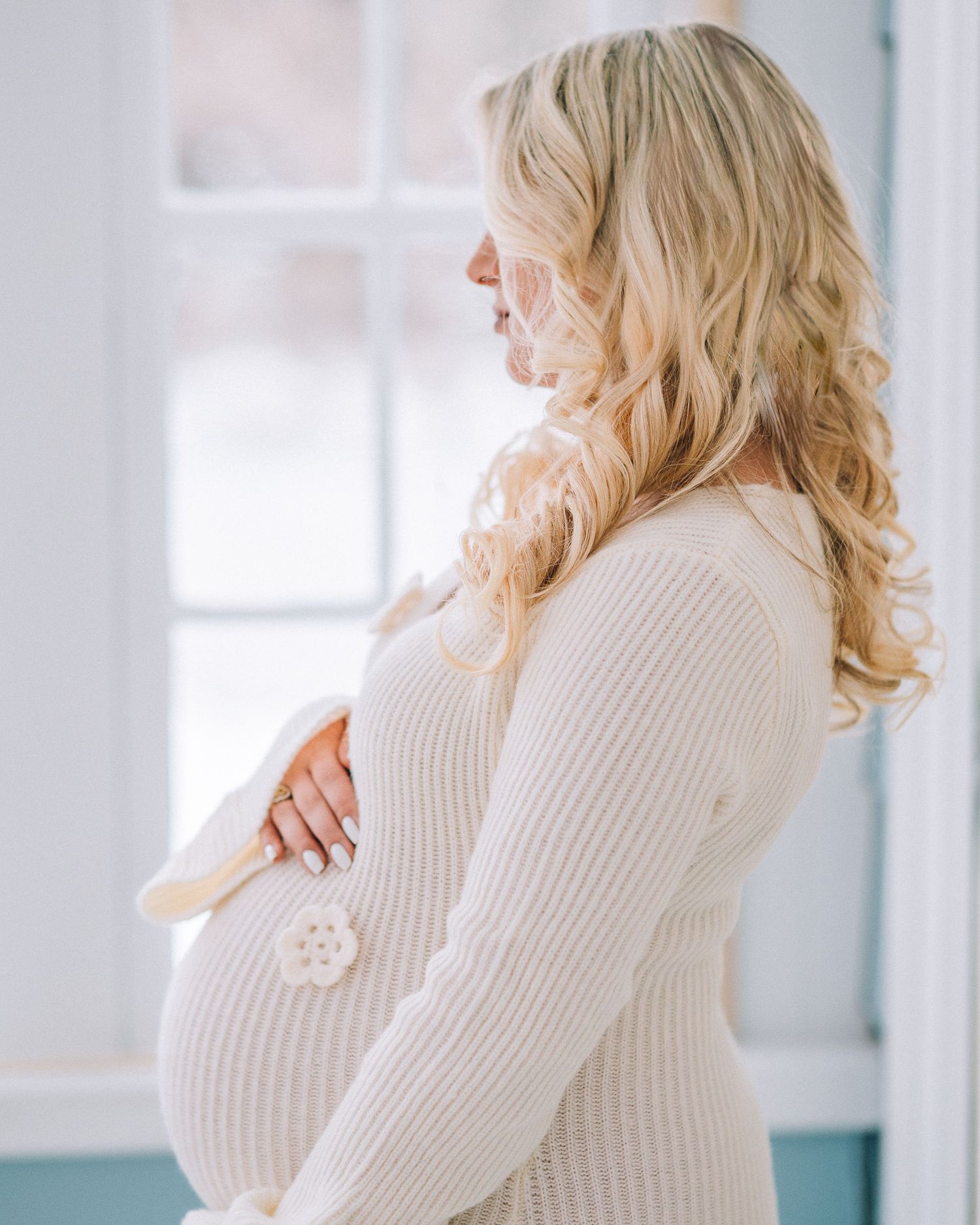 Pregnant woman in cream ribbed sweater stands in profile by window, gently holding her baby bump.