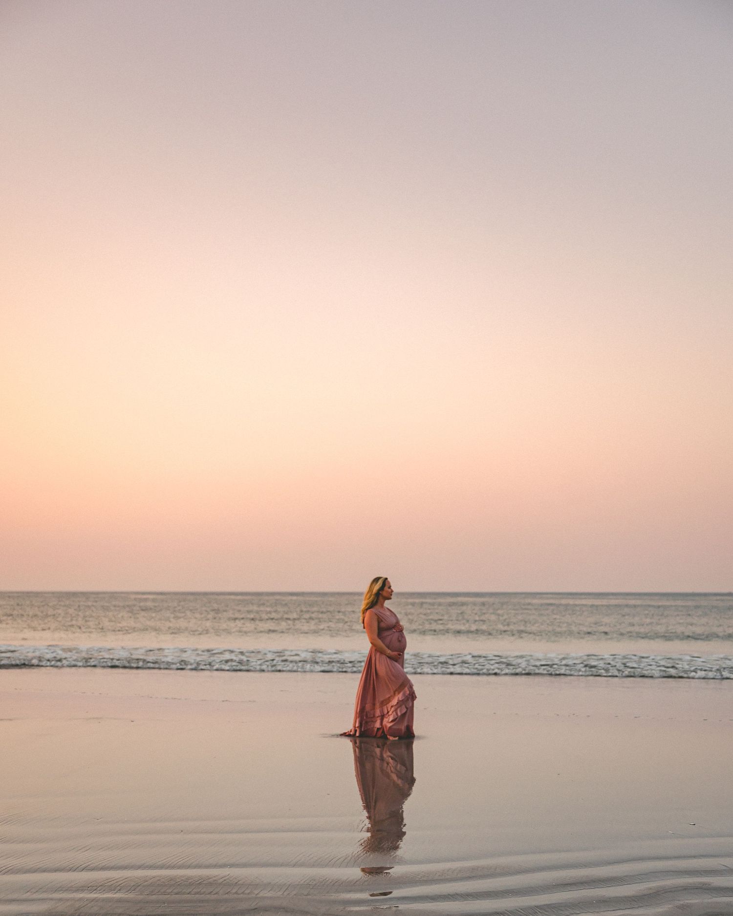 Maternity photo of woman in pink dress standing in shallow ocean water during golden sunset.