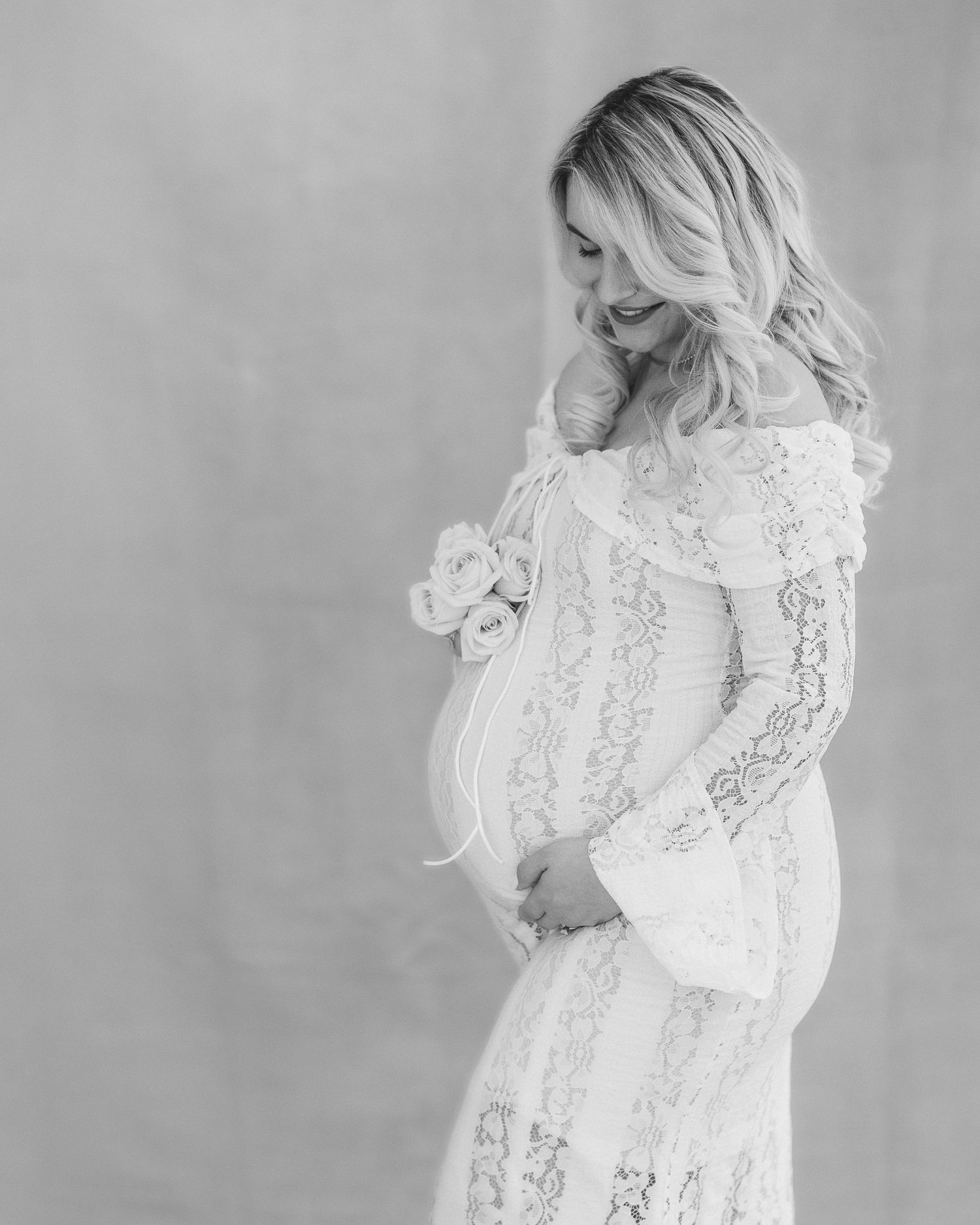 Black and white portrait of an expectant mother in a flowing lace dress smiling while cradling her baby bump.