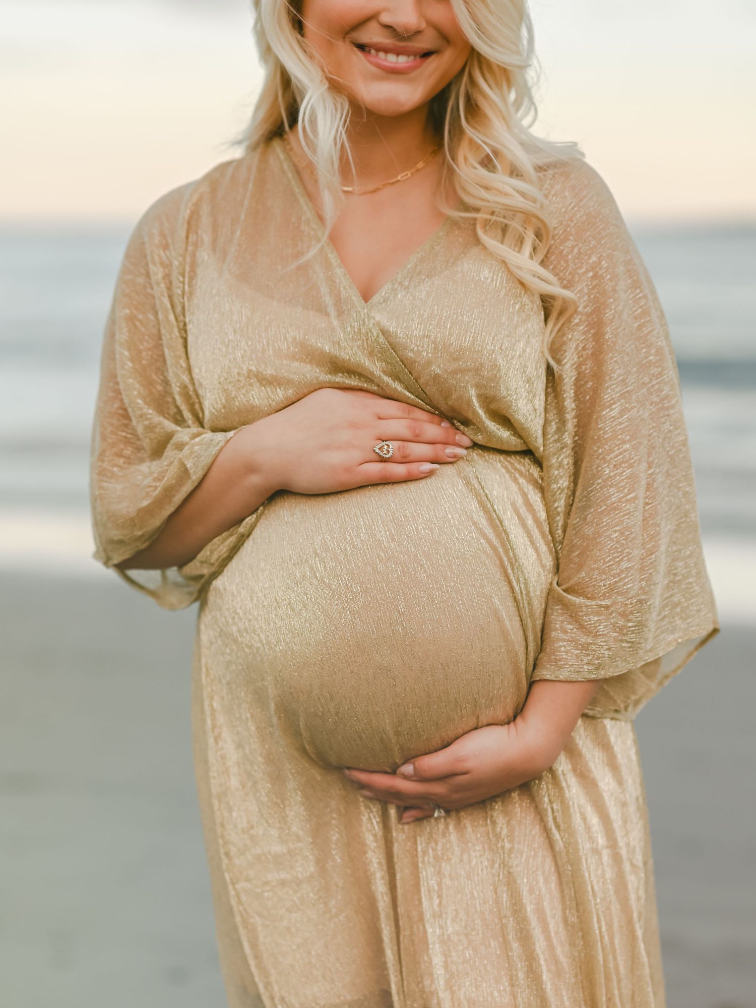 Expecting mother in shimmering gold dress cradles baby bump while standing on beach at sunset.