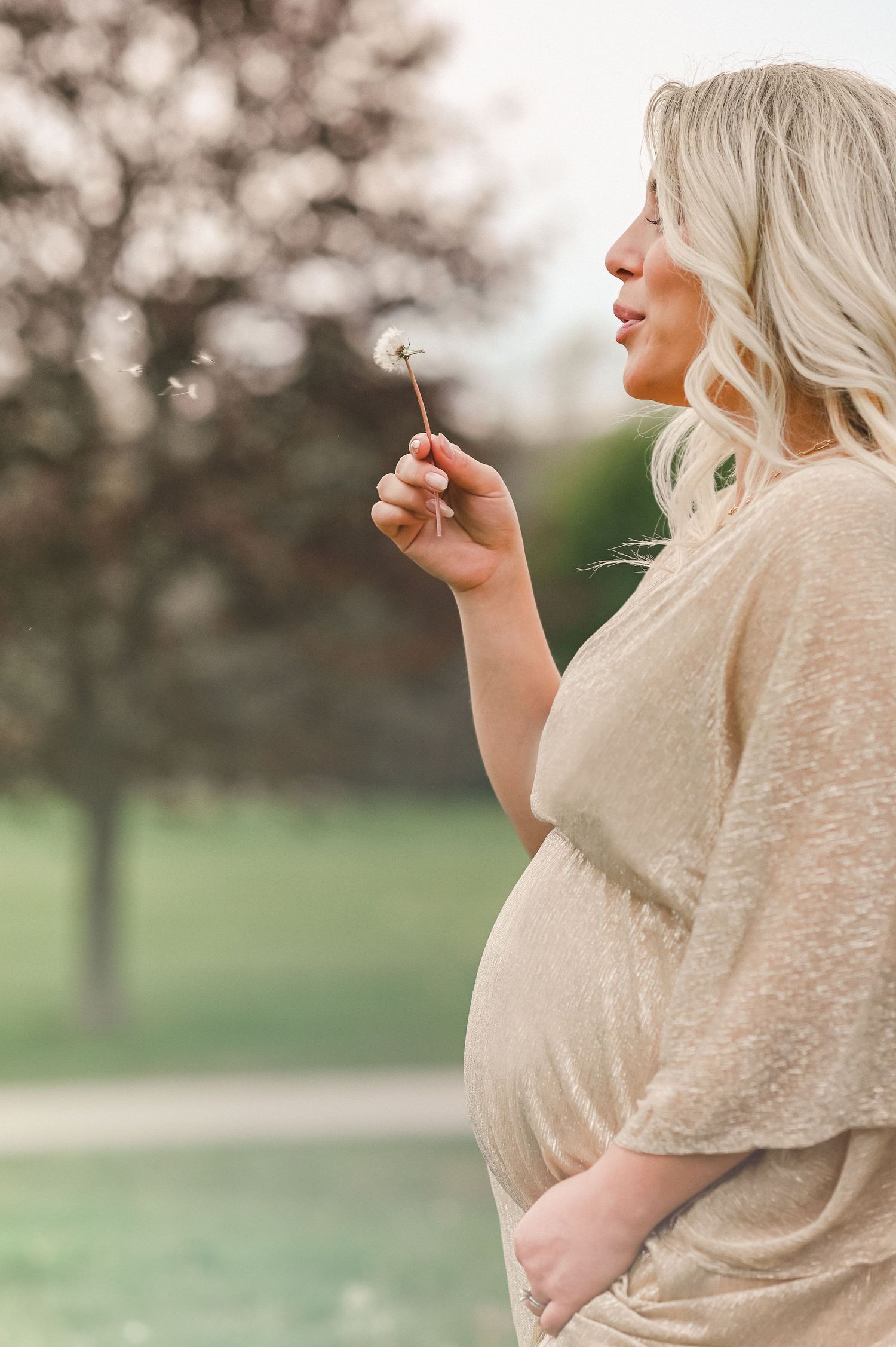 Expectant mother in beige dress holds dandelion while standing in sunlit field during golden hour.