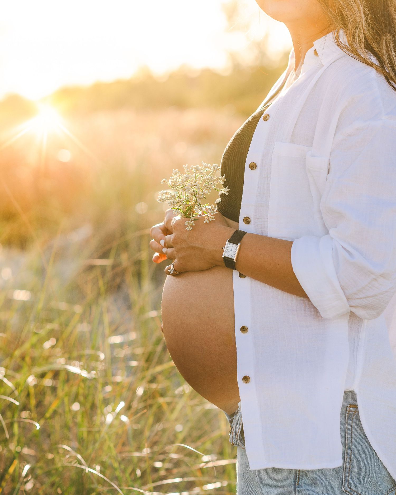 Maternity photo shoot in a sunlit meadow with white flowers as person in white shirt touches their pregnant belly.