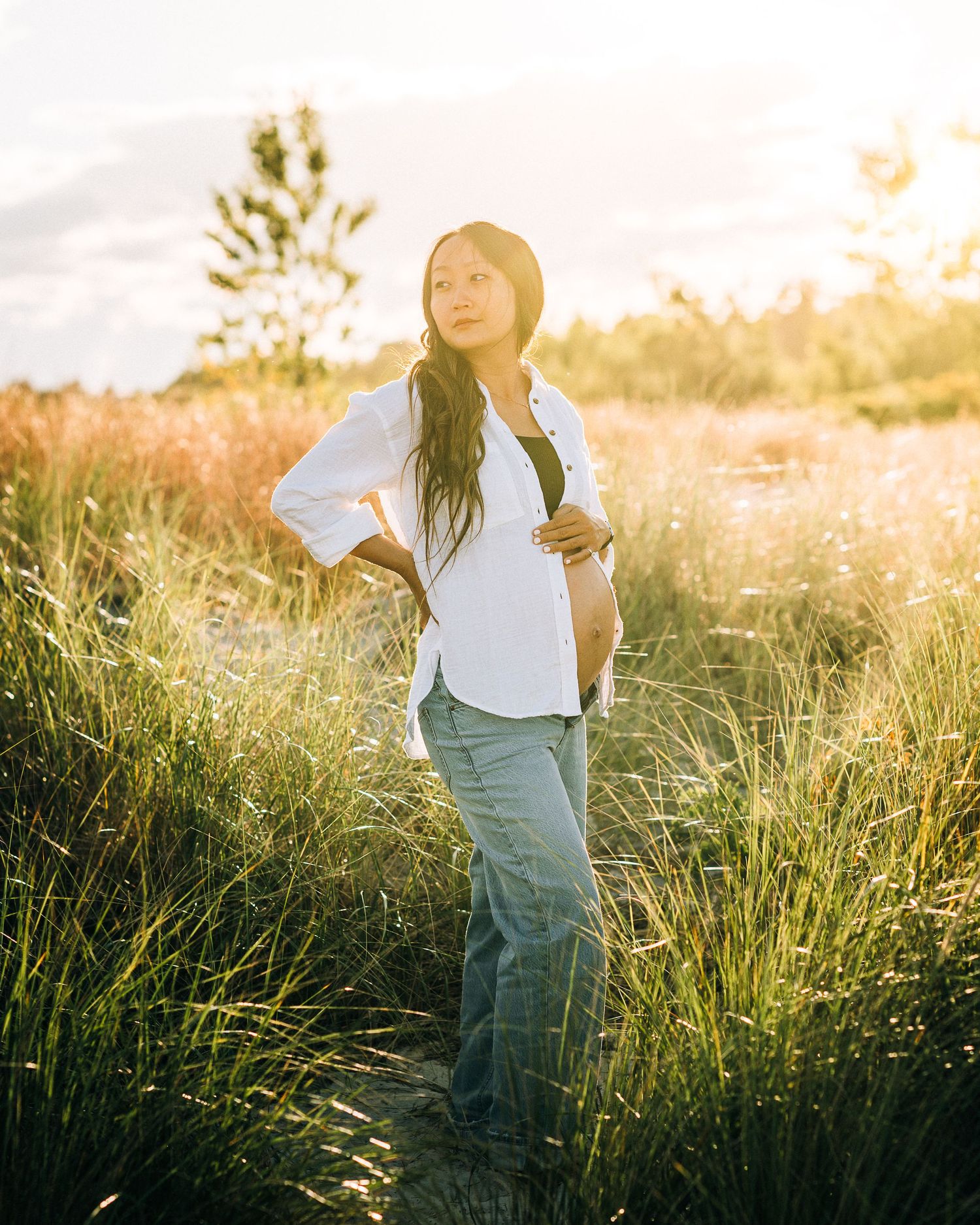 Person in casual attire stands in tall grass field during golden hour sunset with glowing backlight.