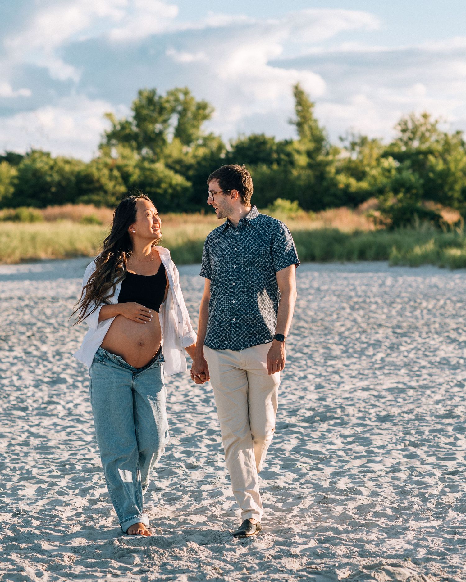 A couple walks together on a sunny beach, sharing a joyful moment against a backdrop of trees and blue sky.