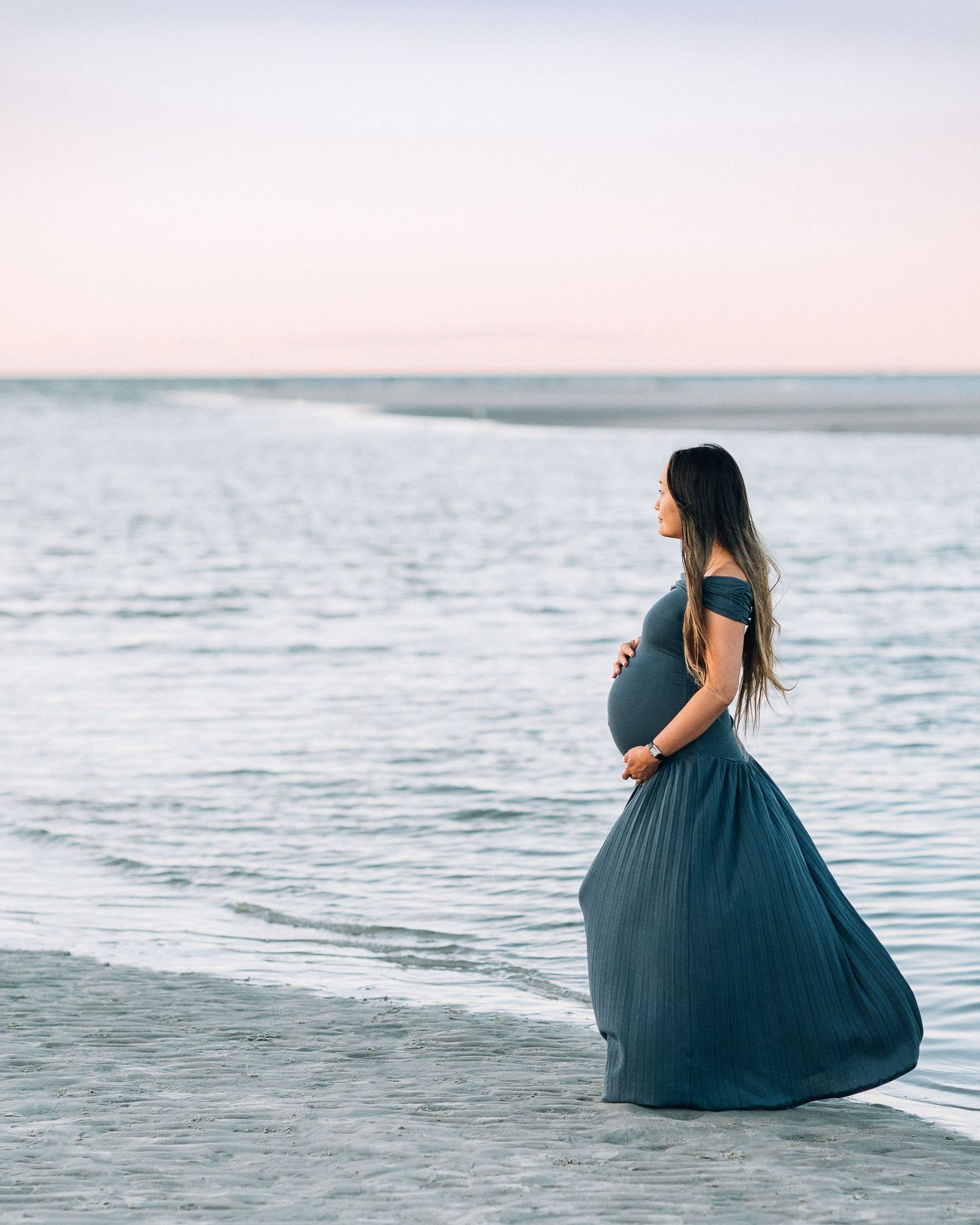 A woman in a flowing navy blue dress sits gracefully on the beach at sunset overlooking the ocean and horizon.