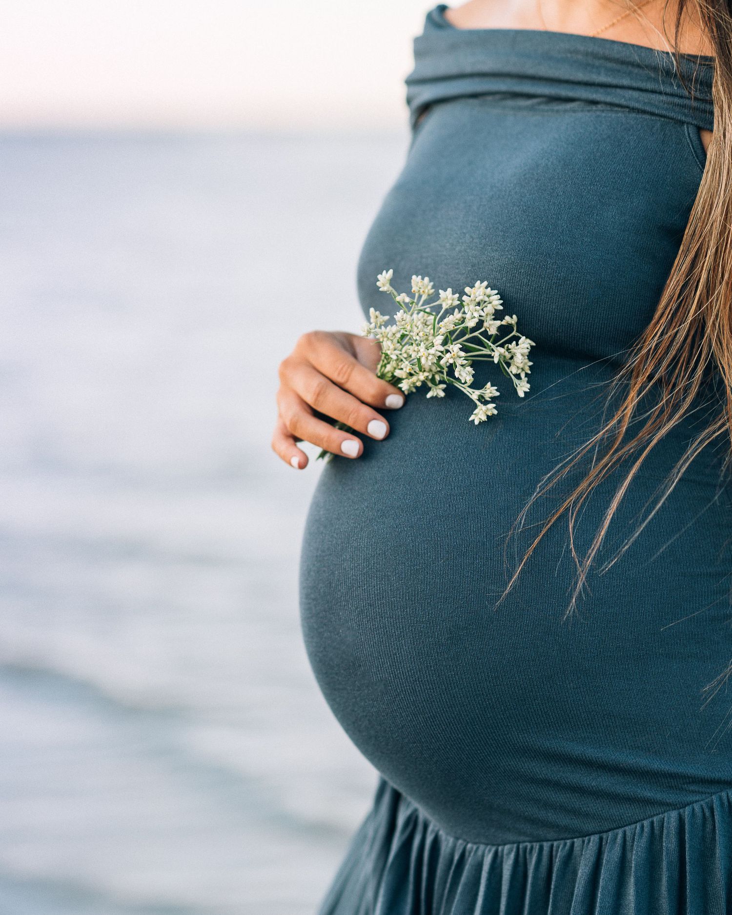 Pregnant figure in a dark teal fitted maternity dress holds delicate white baby's breath flowers near belly in profile view.