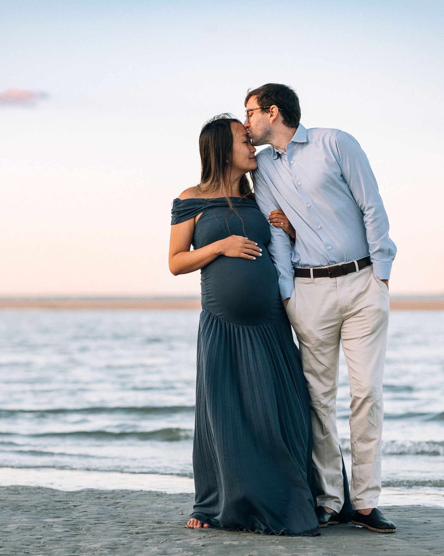 A couple shares a romantic moment on the beach at sunset while expecting a baby.
