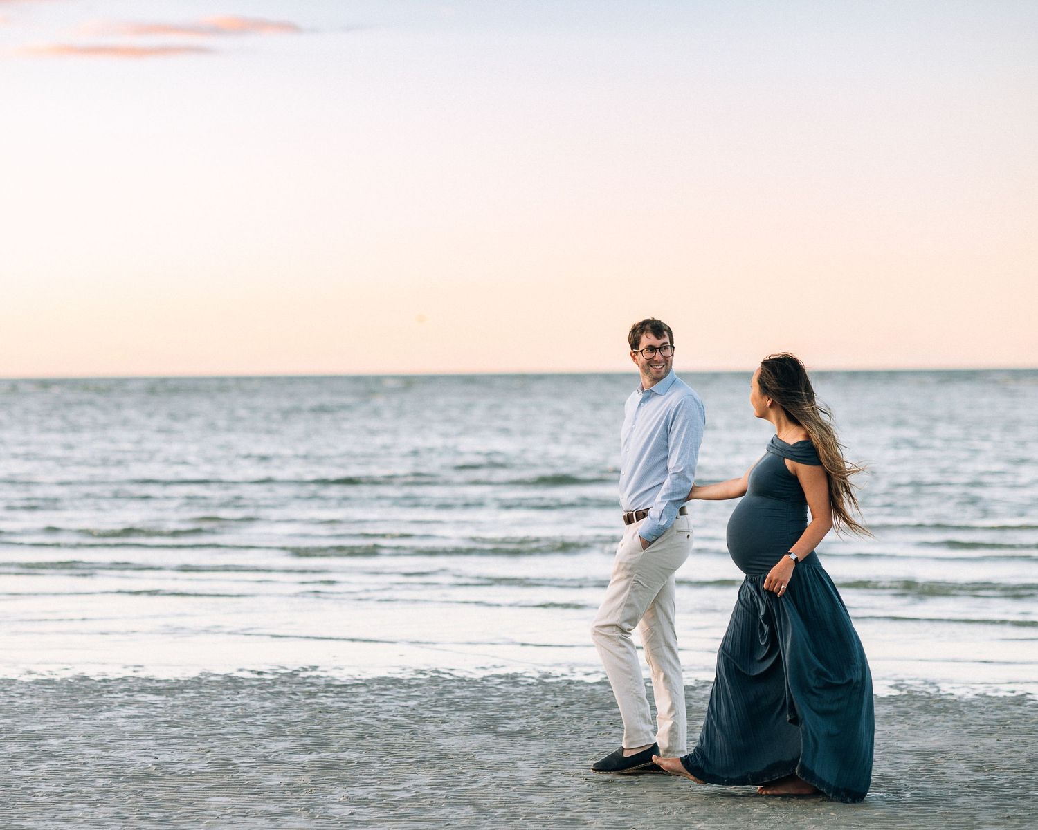 A couple stands together at sunset on a peaceful beach, with the woman wearing a flowing navy blue dress.