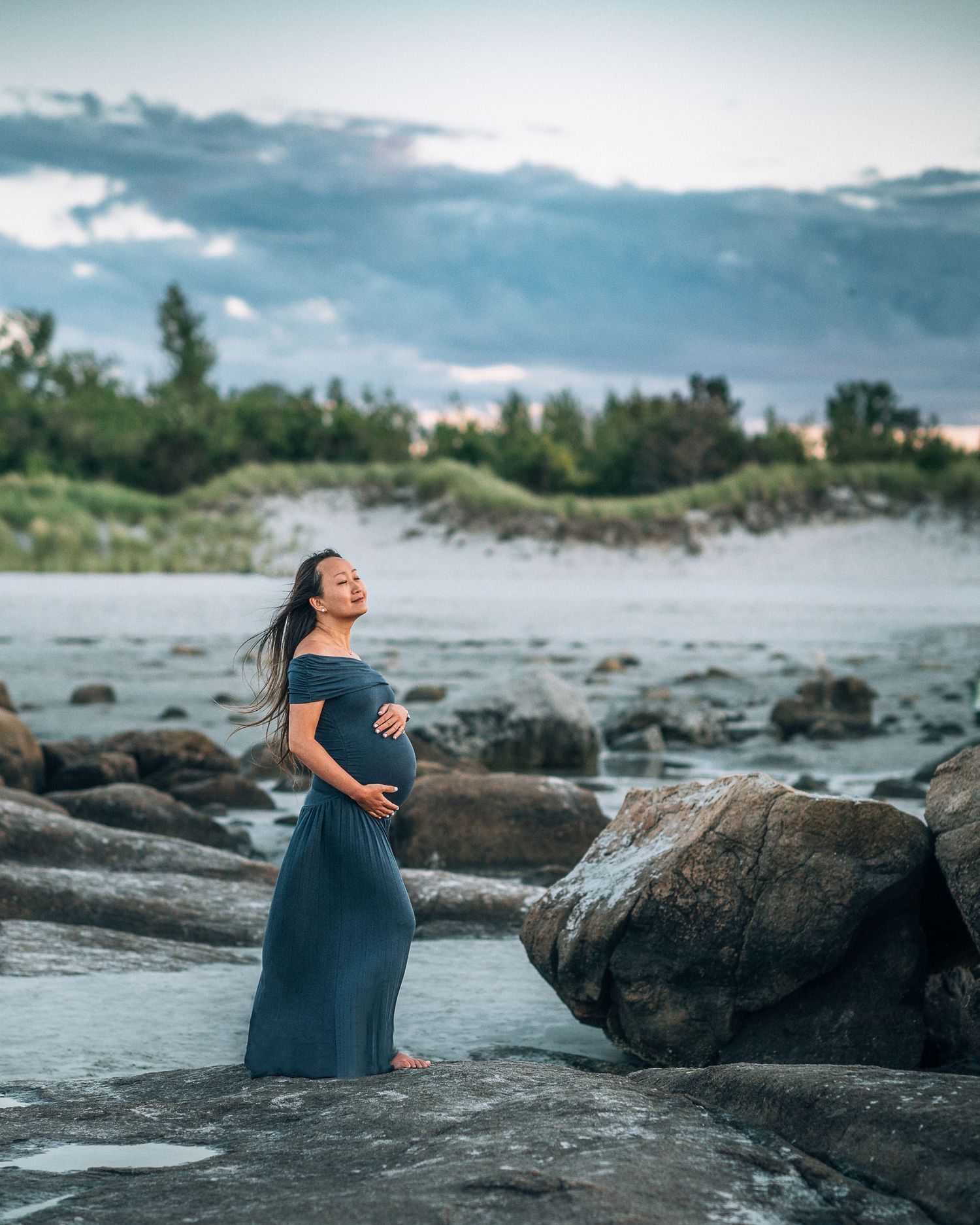 A pregnant woman in a teal dress stands gracefully among rocky coastal scenery during a maternity photoshoot at sunset.