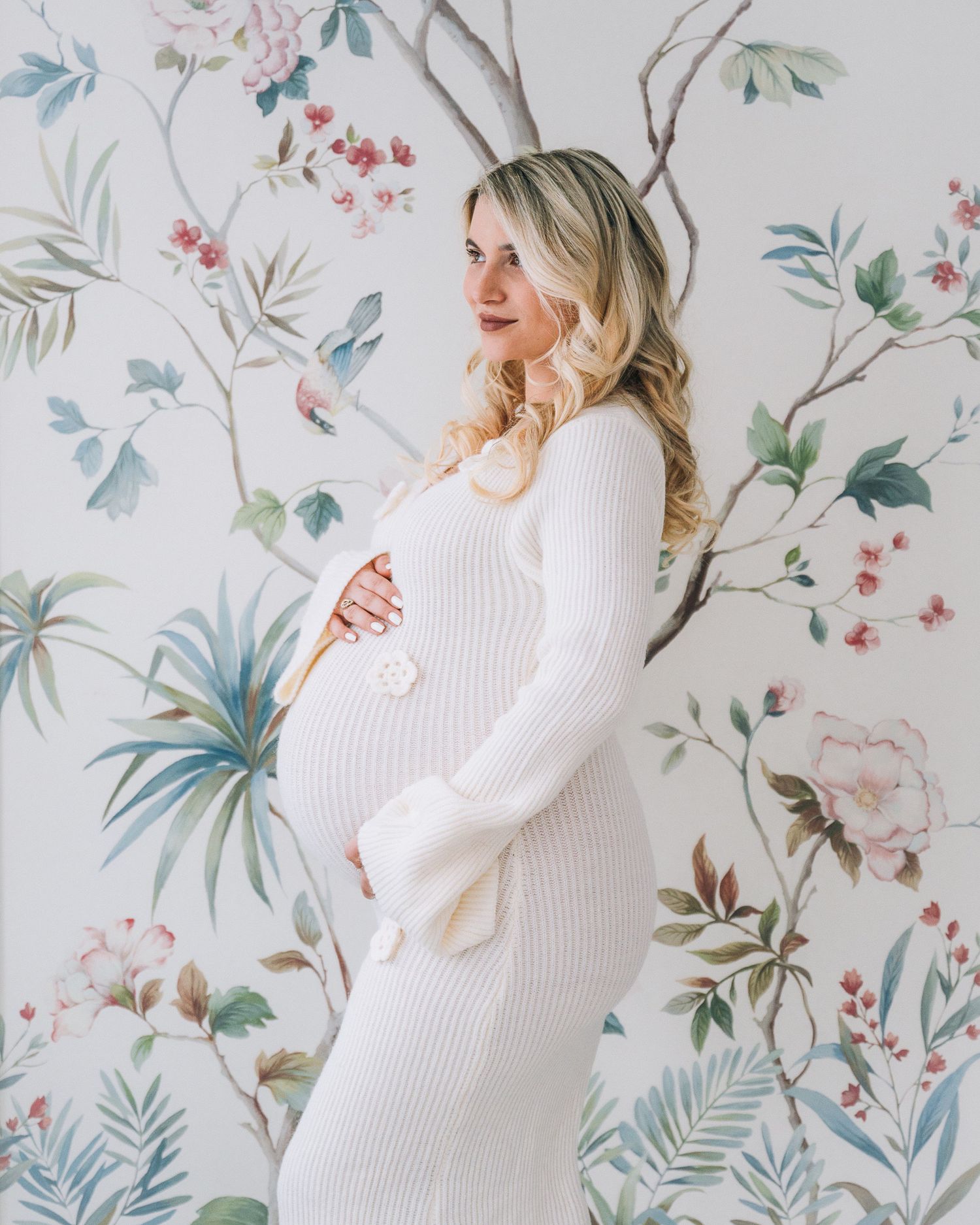 Expectant woman in white sweater dress posing against floral wallpaper with blue and pink botanical design.