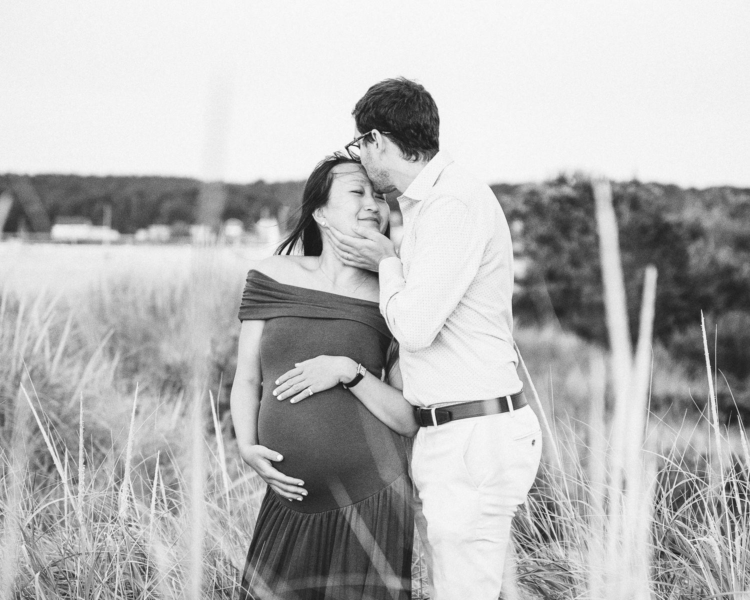 A tender black and white maternity photo of a couple embracing in a sunlit meadow with tall grass.