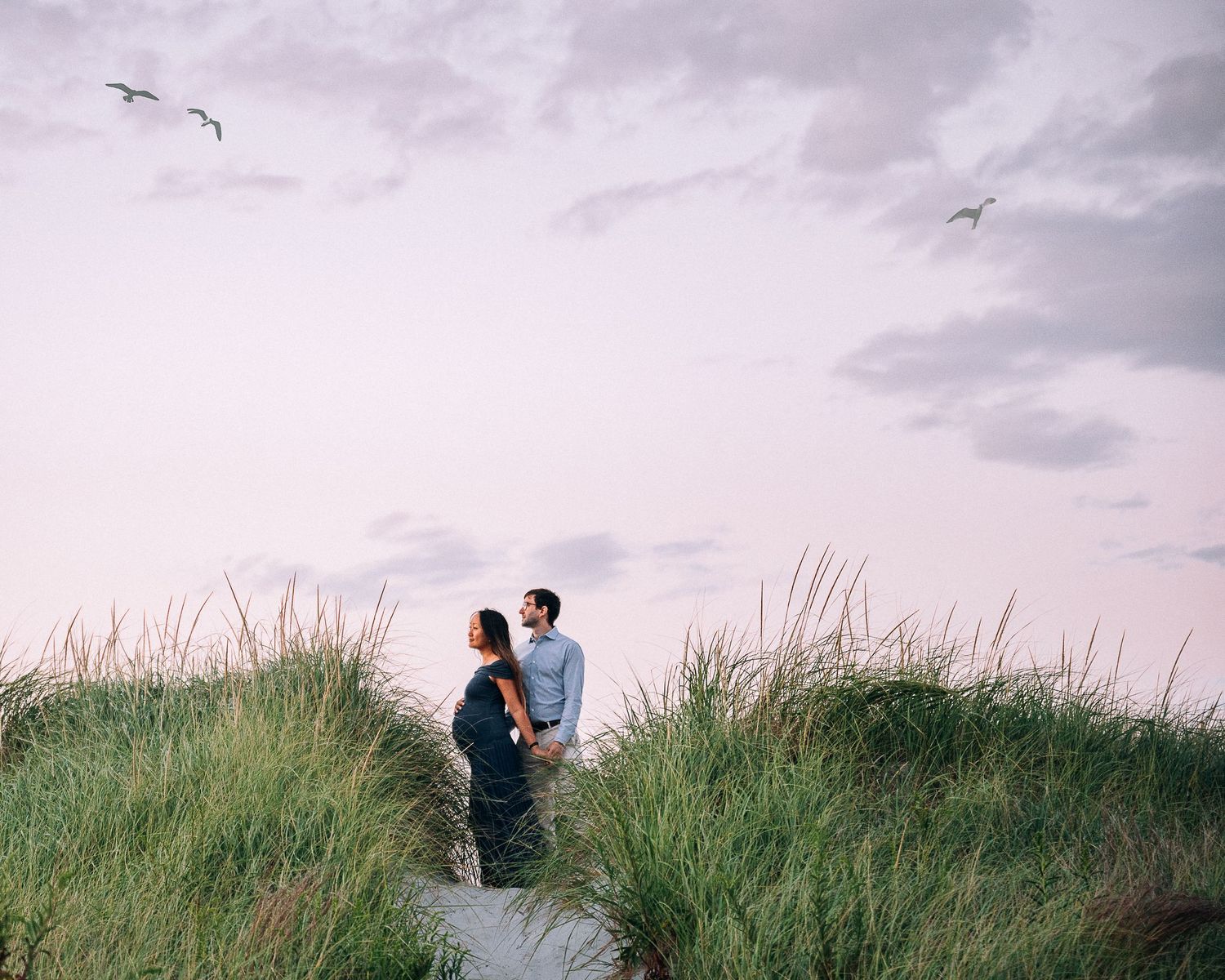 A couple stands amongst tall beach grass on sand dunes as seagulls fly overhead against a pink sunset sky.