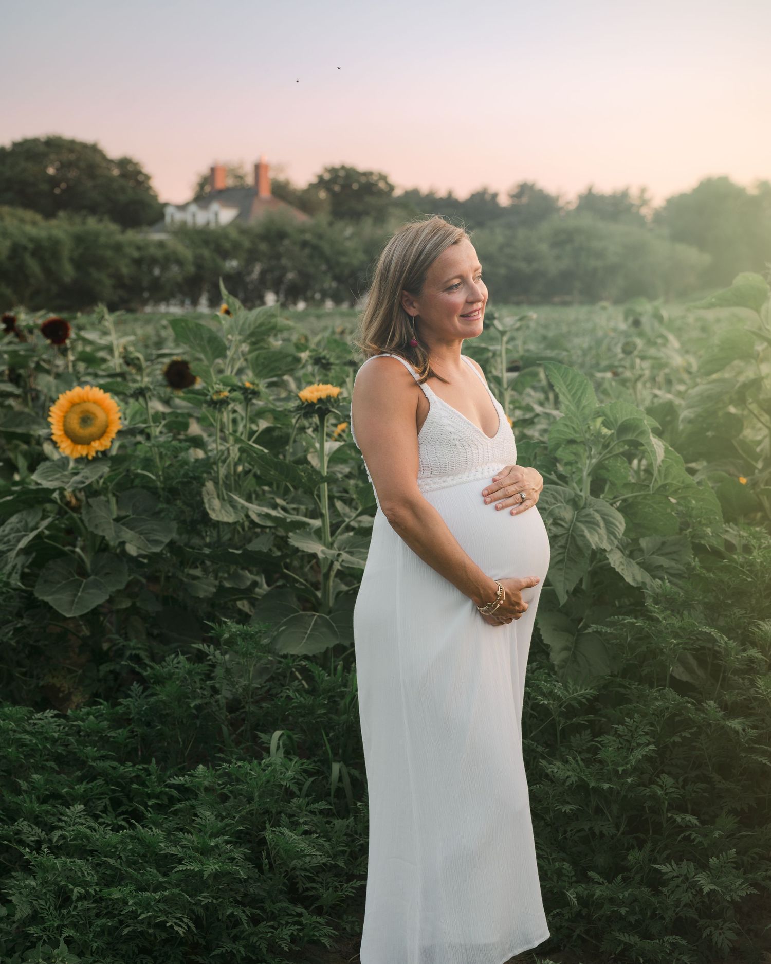 A person in a white dress stands in a sunflower field at sunset, cradling their belly in a maternity photo.