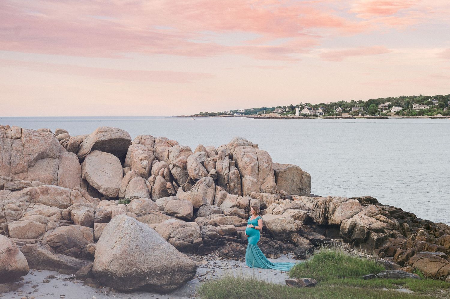 A figure in a flowing turquoise dress poses on rocky coastal shoreline during sunset with houses visible in distance.