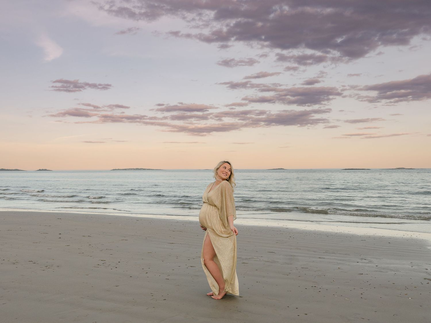 Person in flowy beige dress stands on beach at sunset while waves roll onto shore and clouds paint the sky.