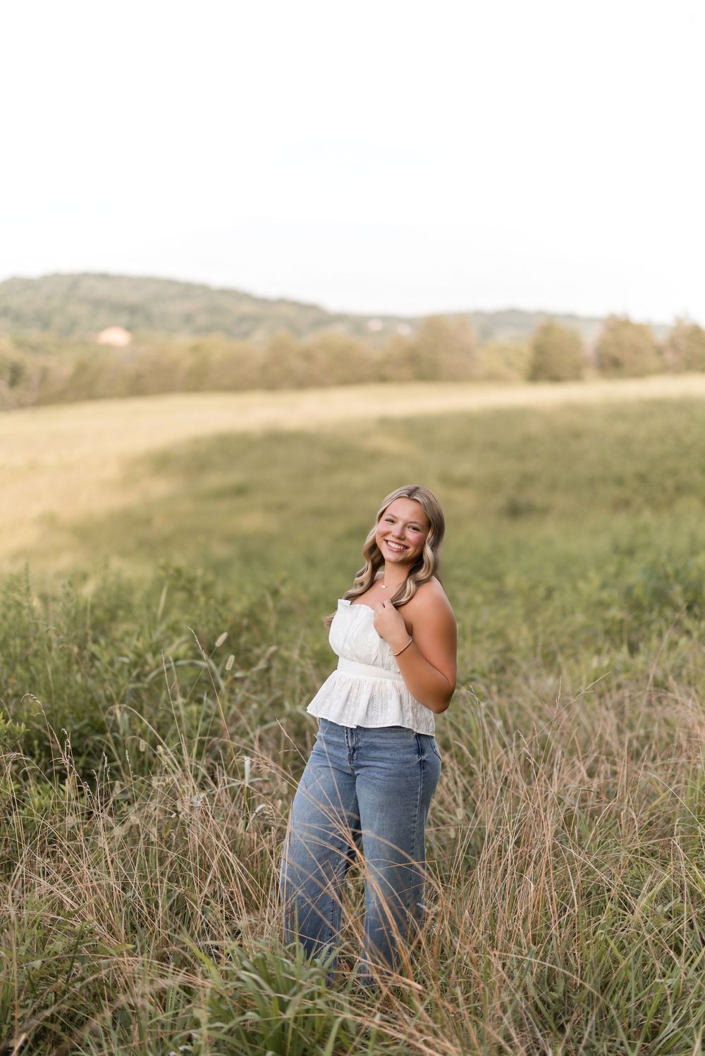 A candid moment capturing someone posing in a grassy meadow field wearing jeans and white top under golden sunlight.