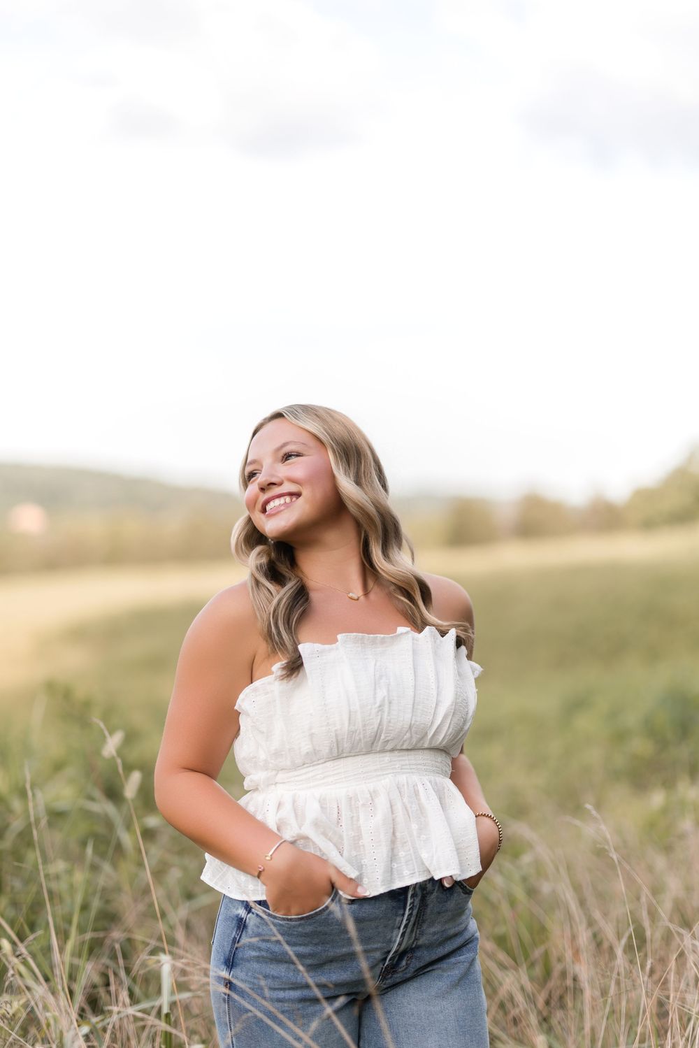 Young woman in white off-shoulder top and jeans smiling outdoors in a sunlit field with tall grass.