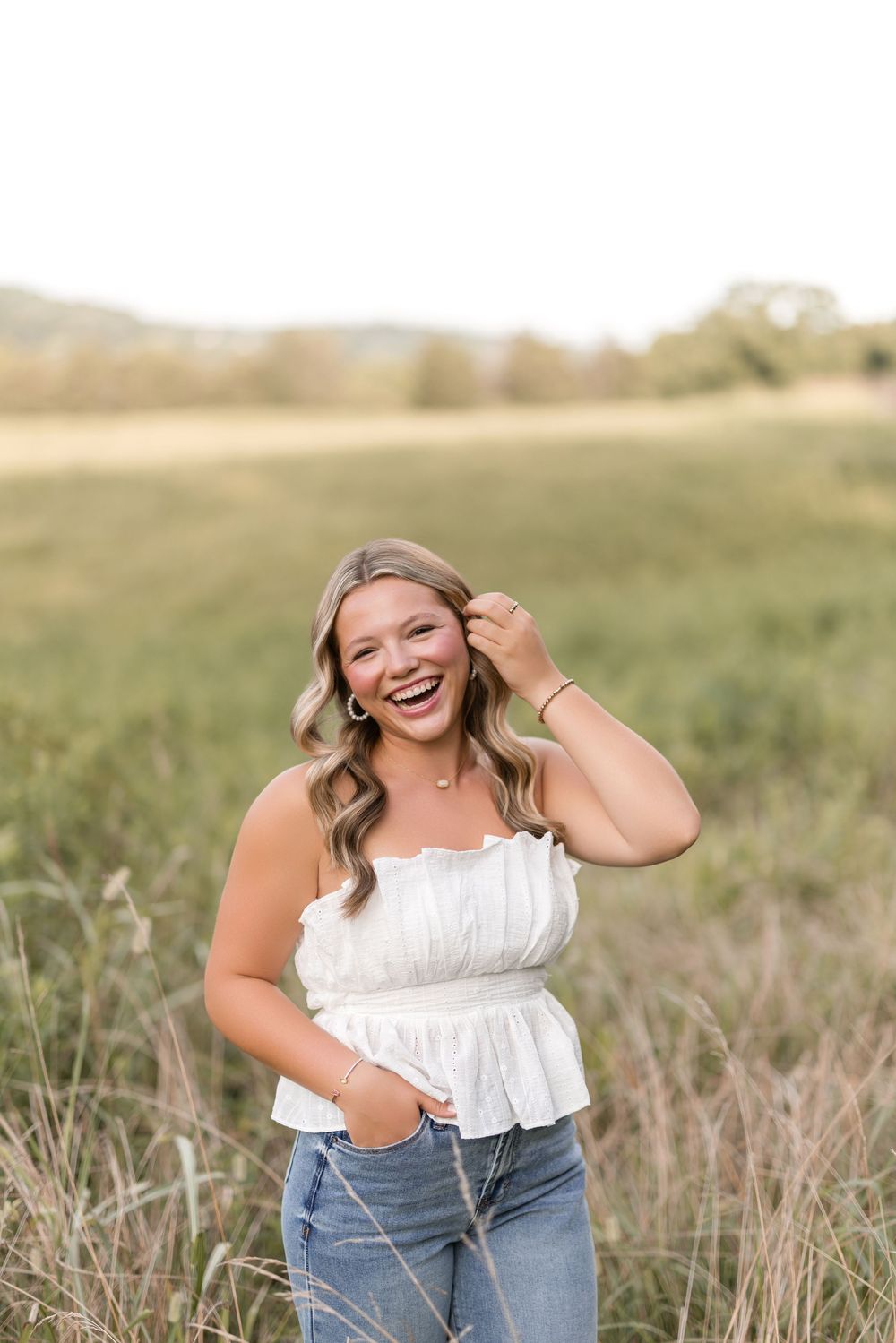 A joyful person wearing a white peplum top and jeans poses in a sunlit field of tall grass.