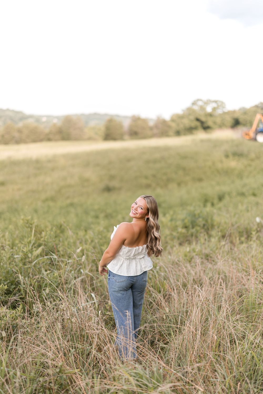 Young woman in white tank top and jeans stands in tall grass field during golden hour in countryside setting.