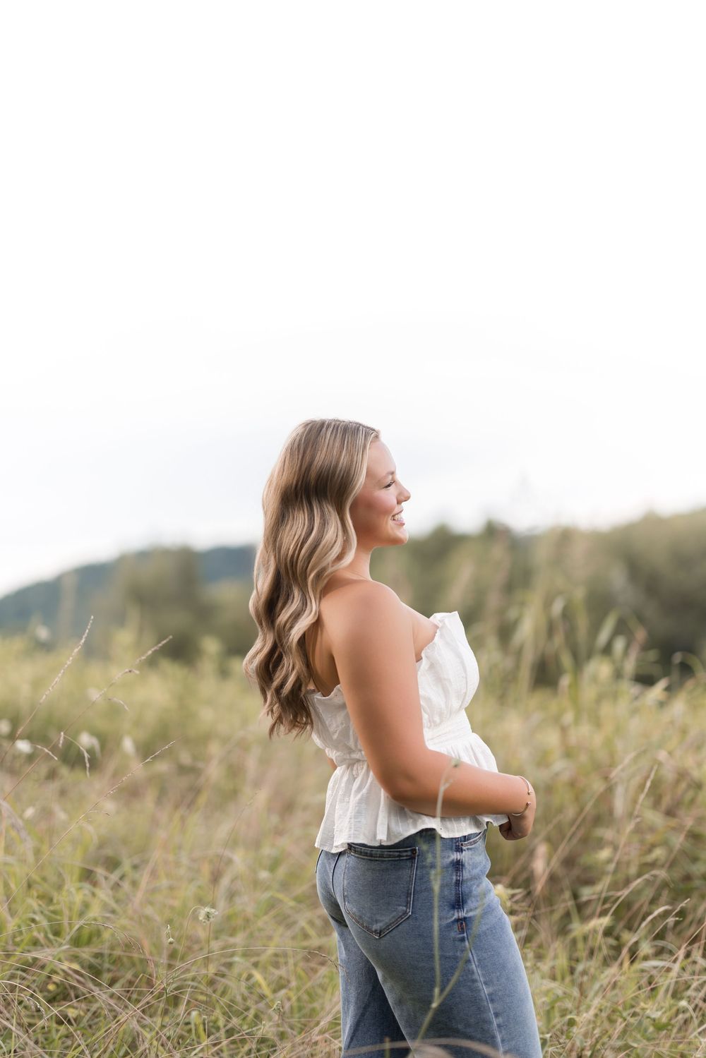 Person in jeans and white top poses joyfully in a sunlit field of tall grass on a warm summer day.