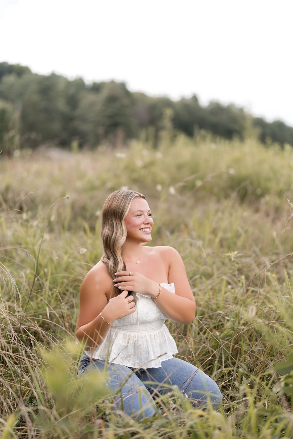 A person wearing a white sleeveless top and jeans sits in a grassy field while laughing joyfully.