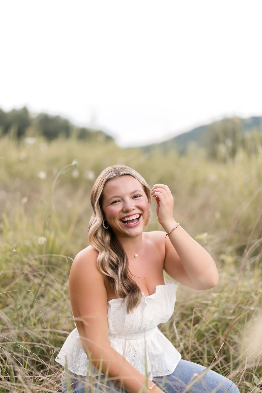 A joyful person wearing a white sleeveless top sits in a meadow of tall grass on a sunny summer day.