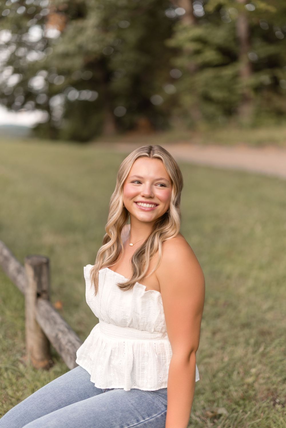 A natural outdoor portrait with someone sitting on a wooden fence wearing a white top and blue jeans on a sunny day.