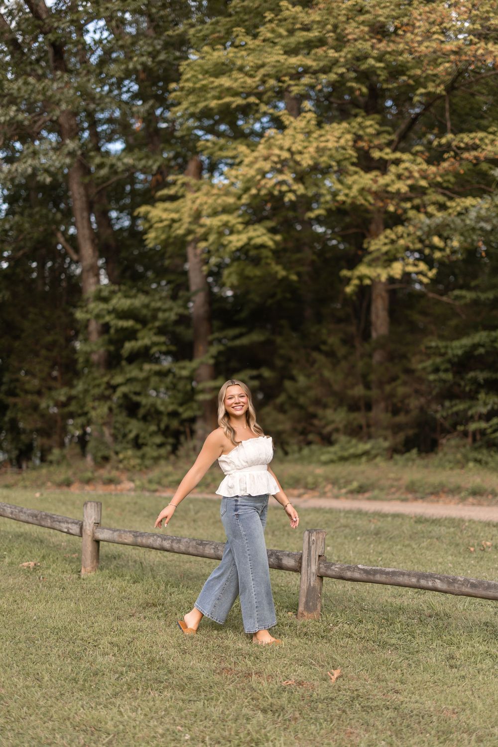 Someone in casual summer attire plays joyfully in a grassy field bordered by wooden fencing and trees.