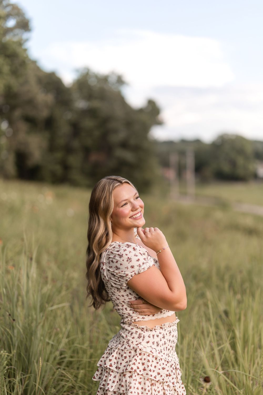 Person in a floral dress laughs joyfully while standing in a sunlit meadow with tall grass.