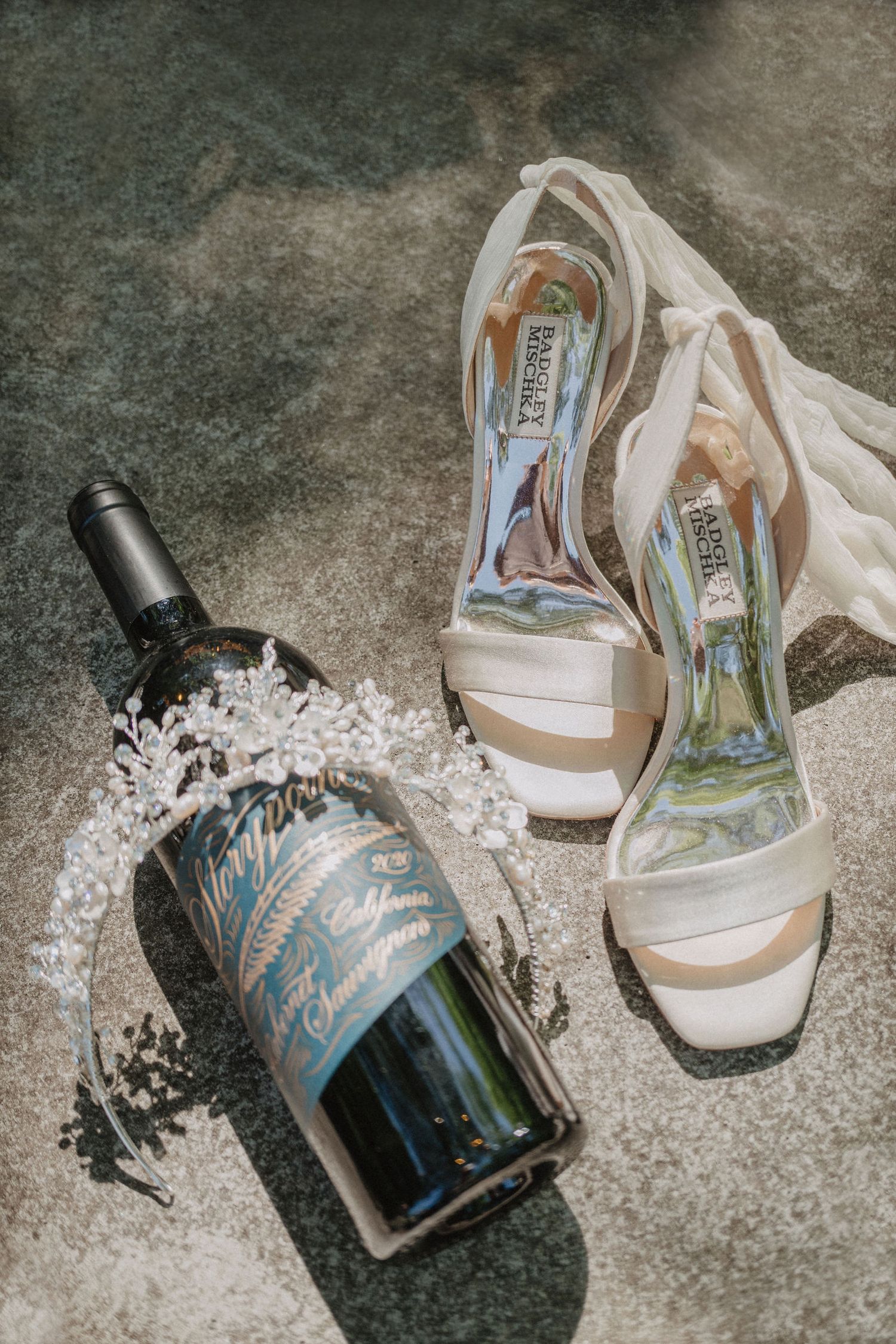 Wedding heels and champagne bottle arranged on concrete surface in natural light.