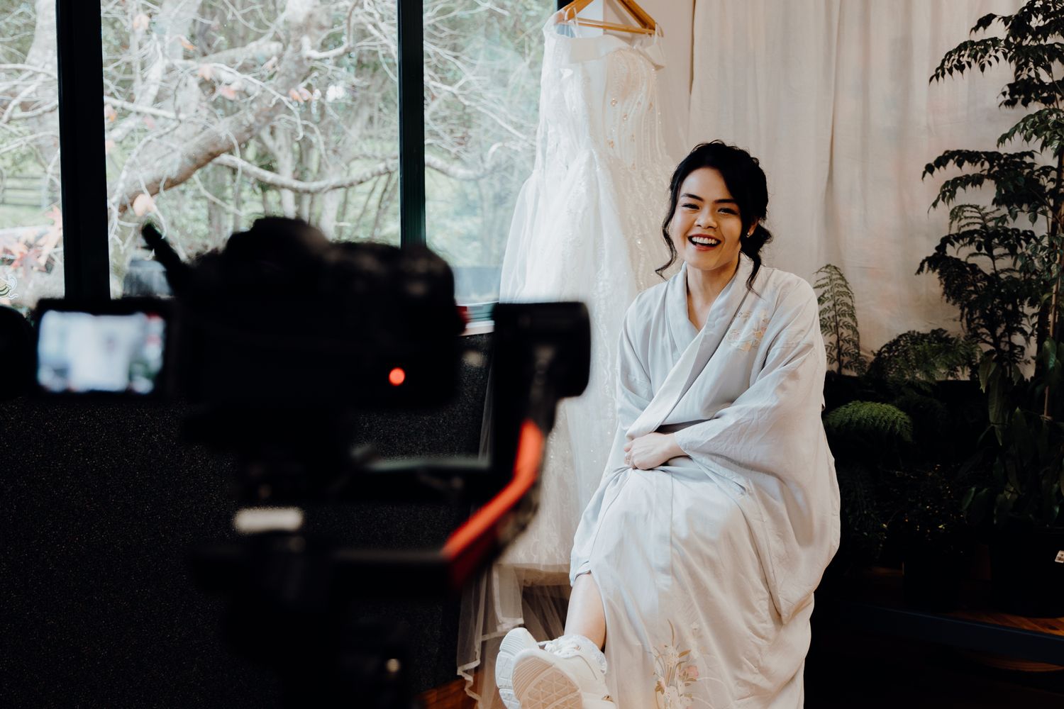 Camera recording wedding preparation scene with person in white robe seated by window.