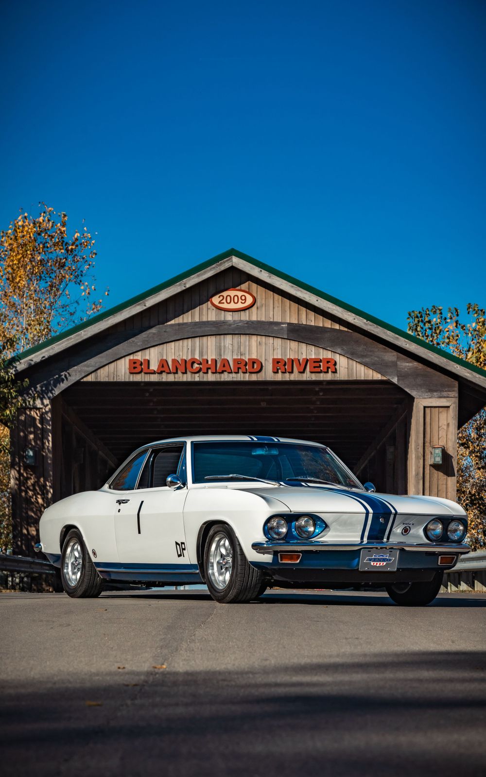 White classic car with blue racing stripes parked in front of wooden Blanchard River building.