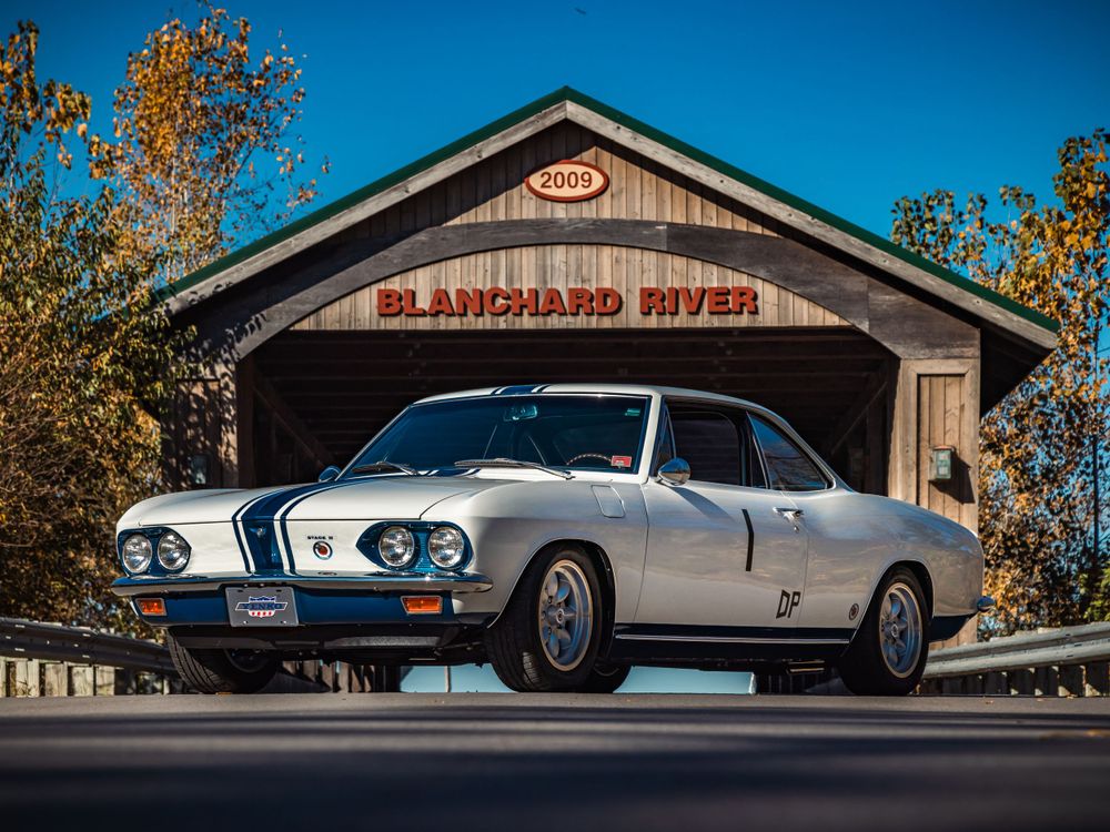 Vintage white sports coupe with racing stripes positioned in front of rustic covered bridge entrance.