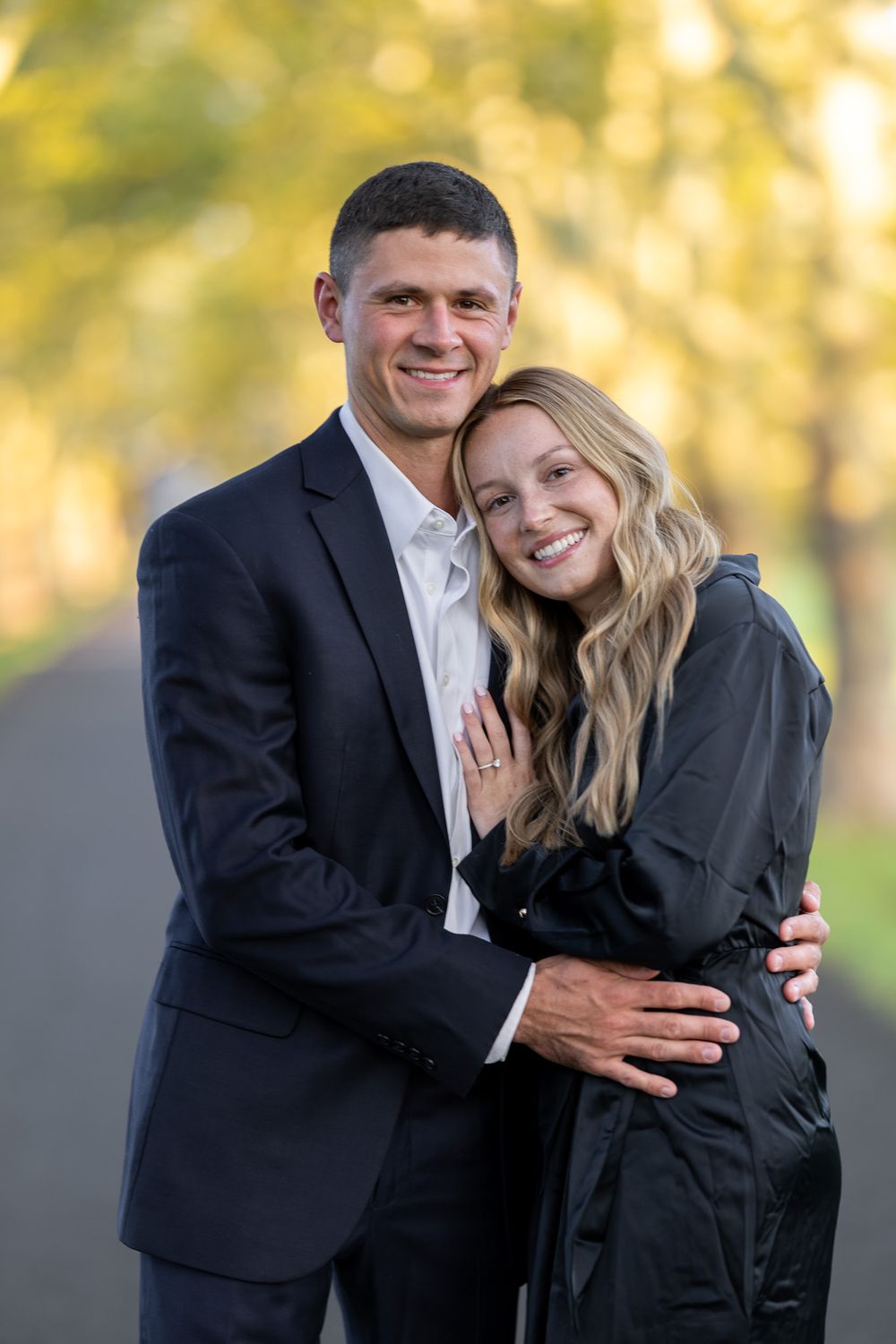 Happy couple in formal attire showing engagement ring on leafy path.