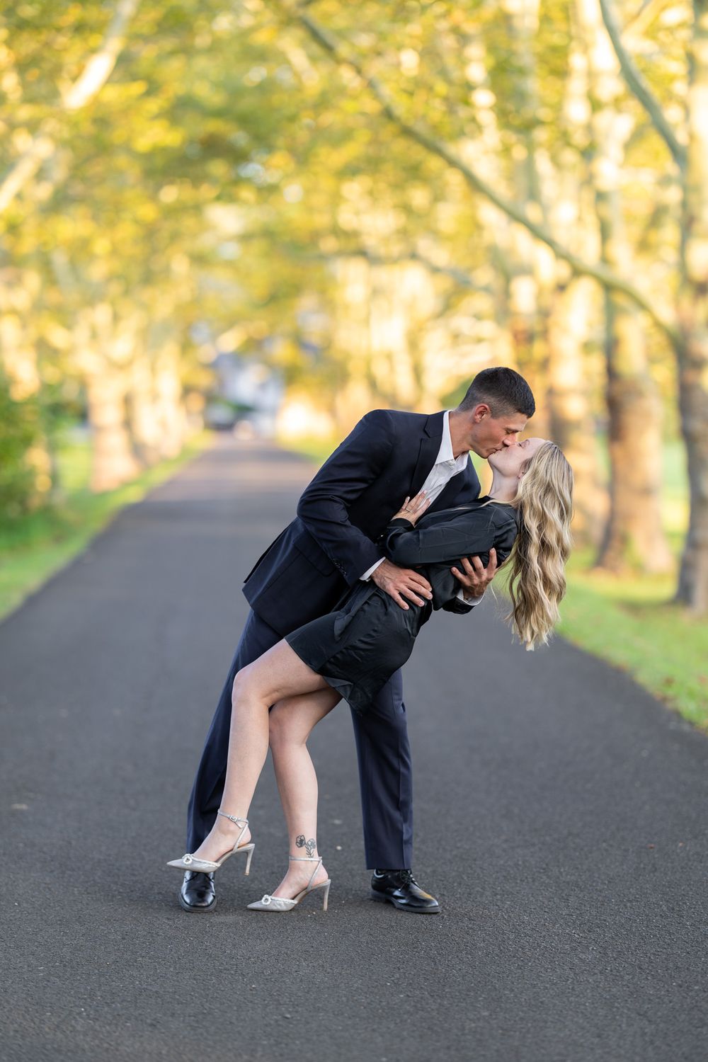 Romantic dip kiss between couple in formal wear on wooded path.