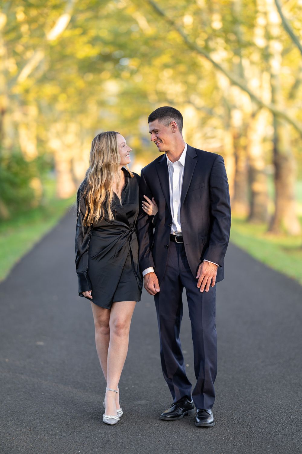 Couple in formal attire walking lovingly together on a tree-lined path during golden hour.
