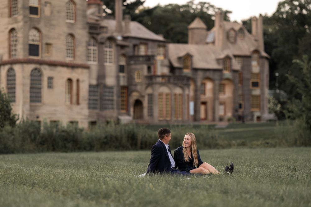 Couple sitting on grass with historic stone castle architecture in background.