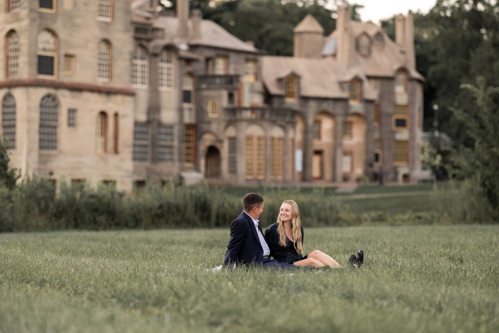 A couple sits on grass in front of a grand historic castle building during a romantic photoshoot.