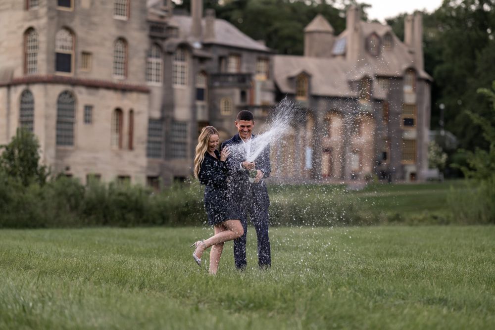 A couple playfully celebrates with champagne spray on castle grounds.