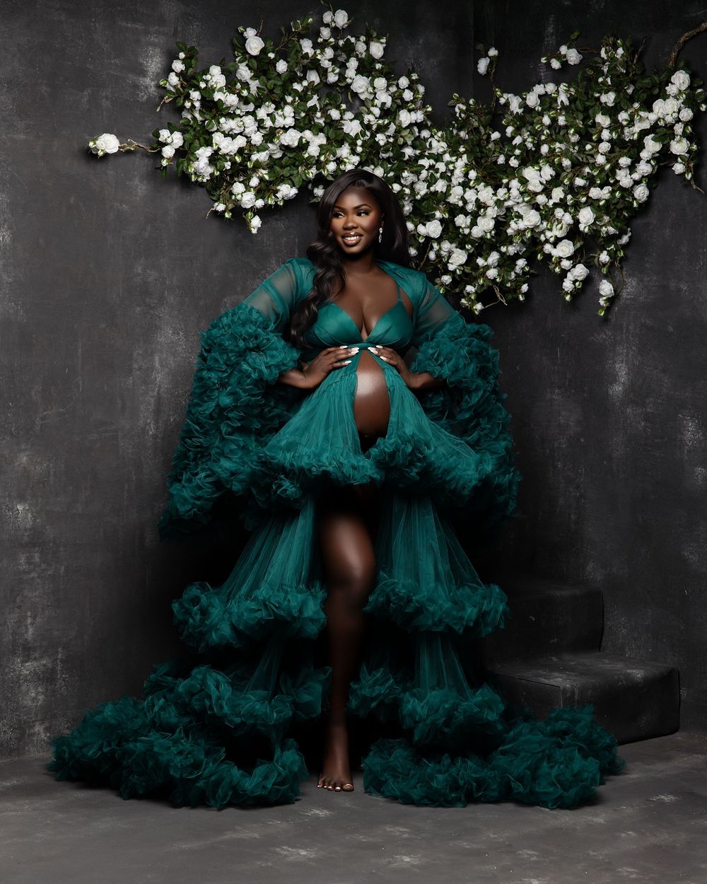 A glamorous portrait in emerald green ruffled tulle gown against a dark backdrop with white floral decorations.