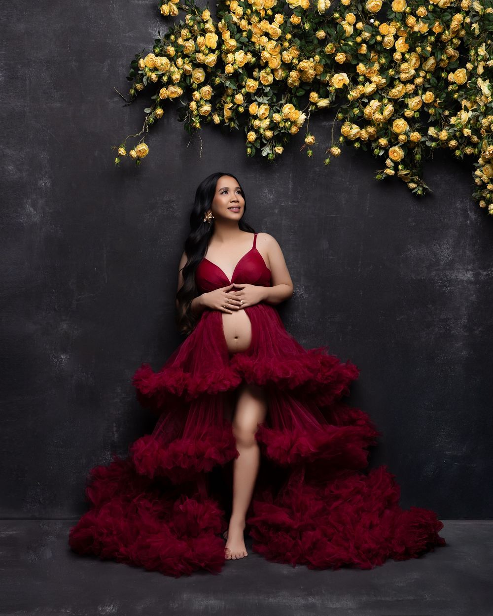 A maternity photo against a dark wall with trailing yellow flowers features a flowing burgundy tulle gown.