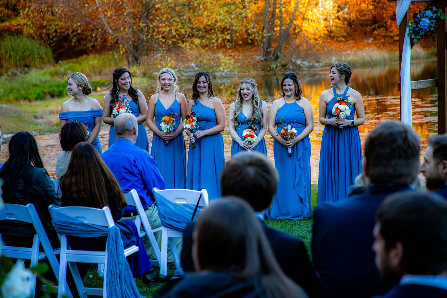 Wedding party in matching bright blue dresses stands outdoors during autumn sunset ceremony.