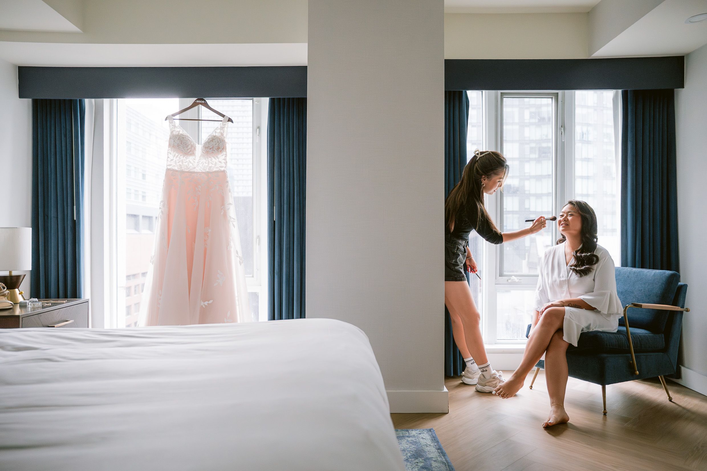 In a bright hotel room with blue curtains, someone gets their makeup done while a blush pink dress hangs by the window.