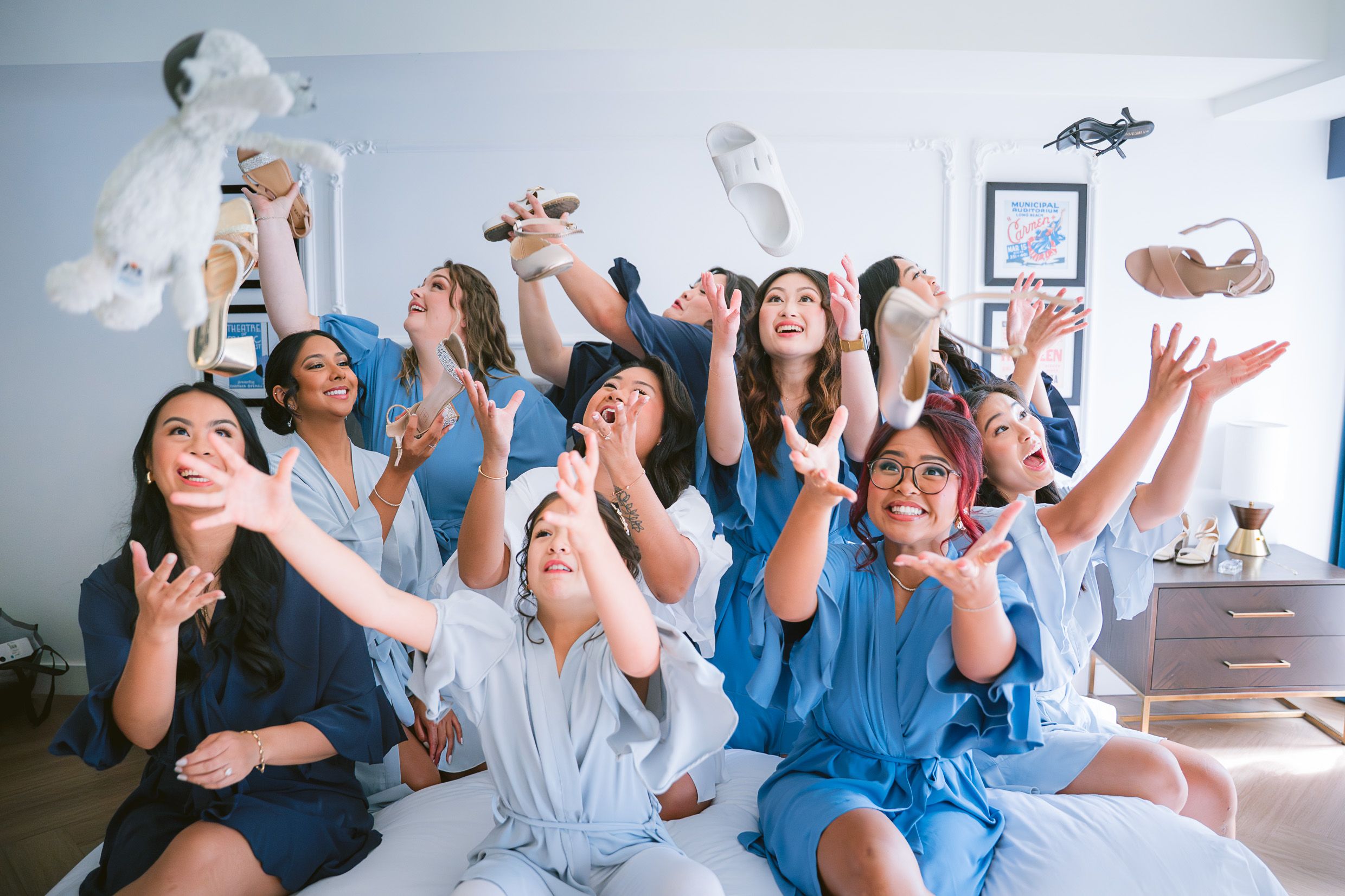 A group of excited friends in matching blue outfits toss flowers and white fabric while celebrating together in a sunlit room.