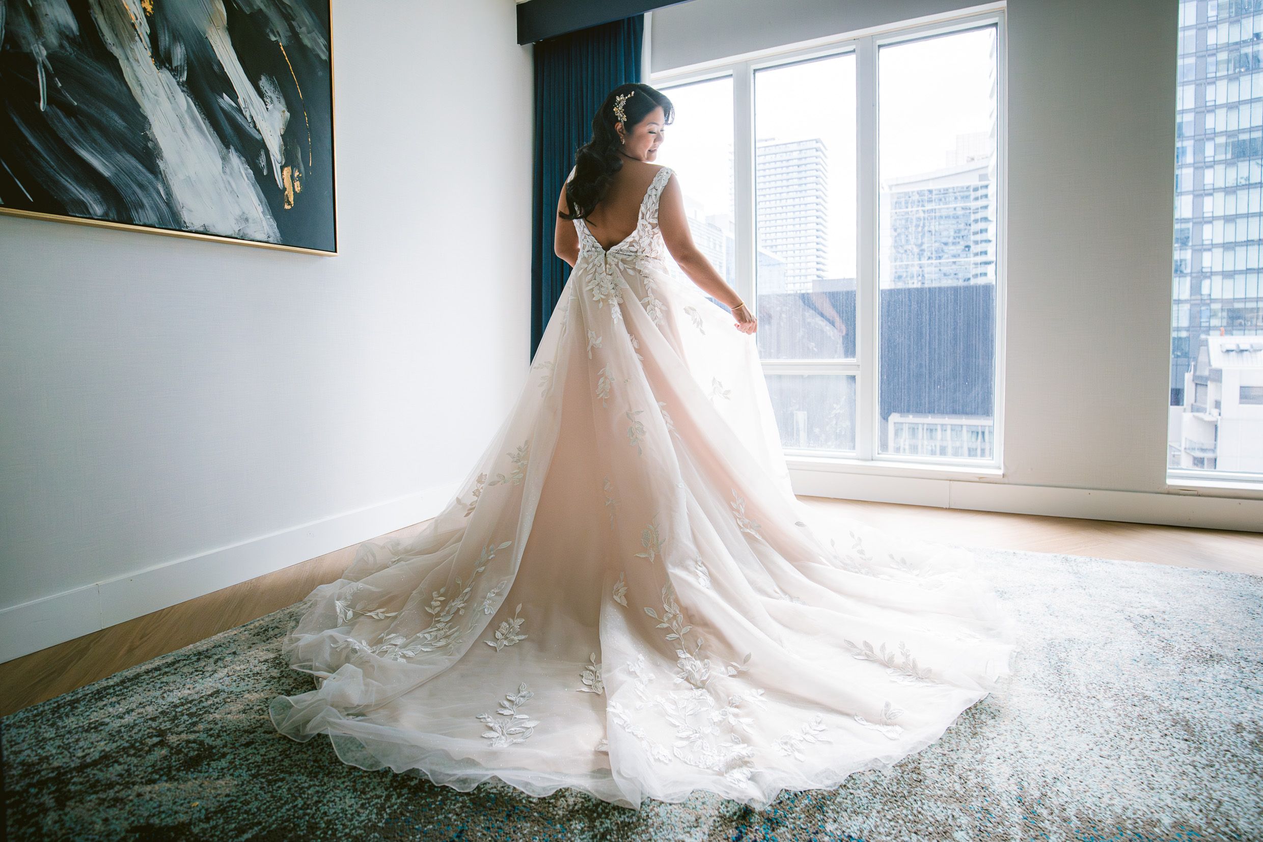 A flowing white wedding gown with a dramatic train is showcased in a modern room with city views.