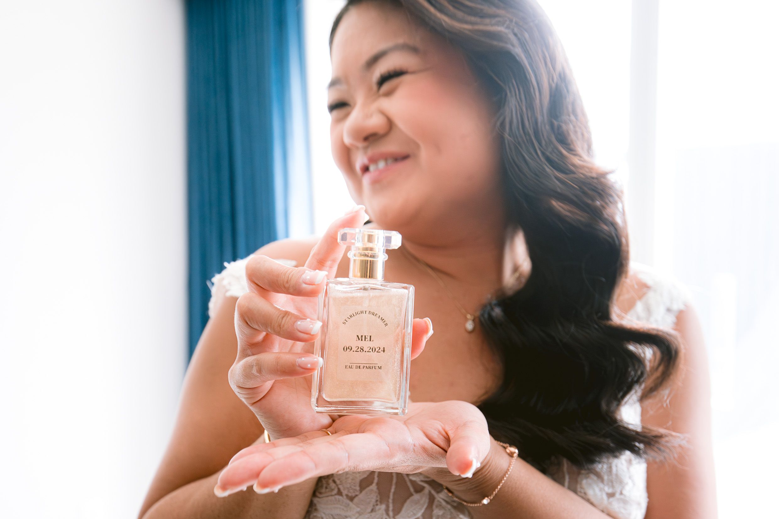 A person holding a delicate glass perfume bottle while smiling warmly in a room with blue curtains.
