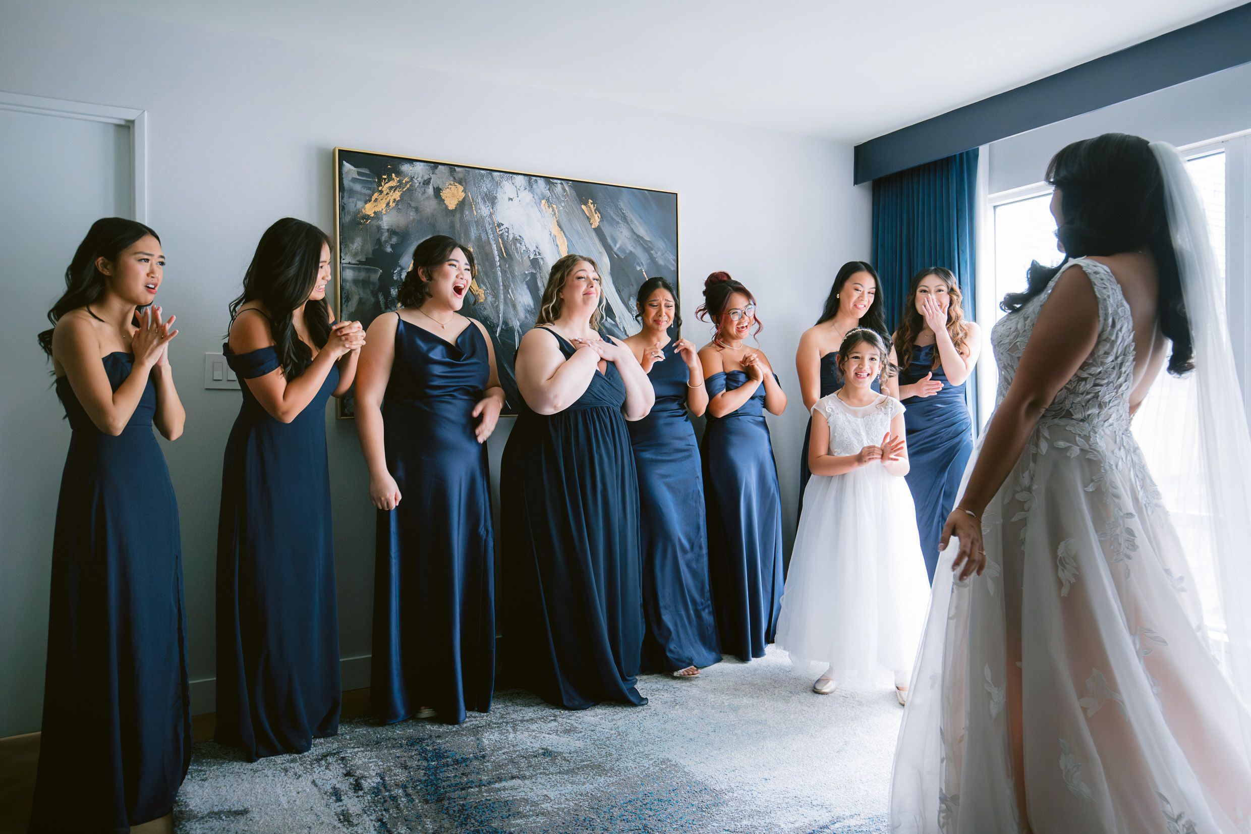 Bridesmaids in matching navy blue dresses react joyfully upon seeing the bride in her wedding gown.