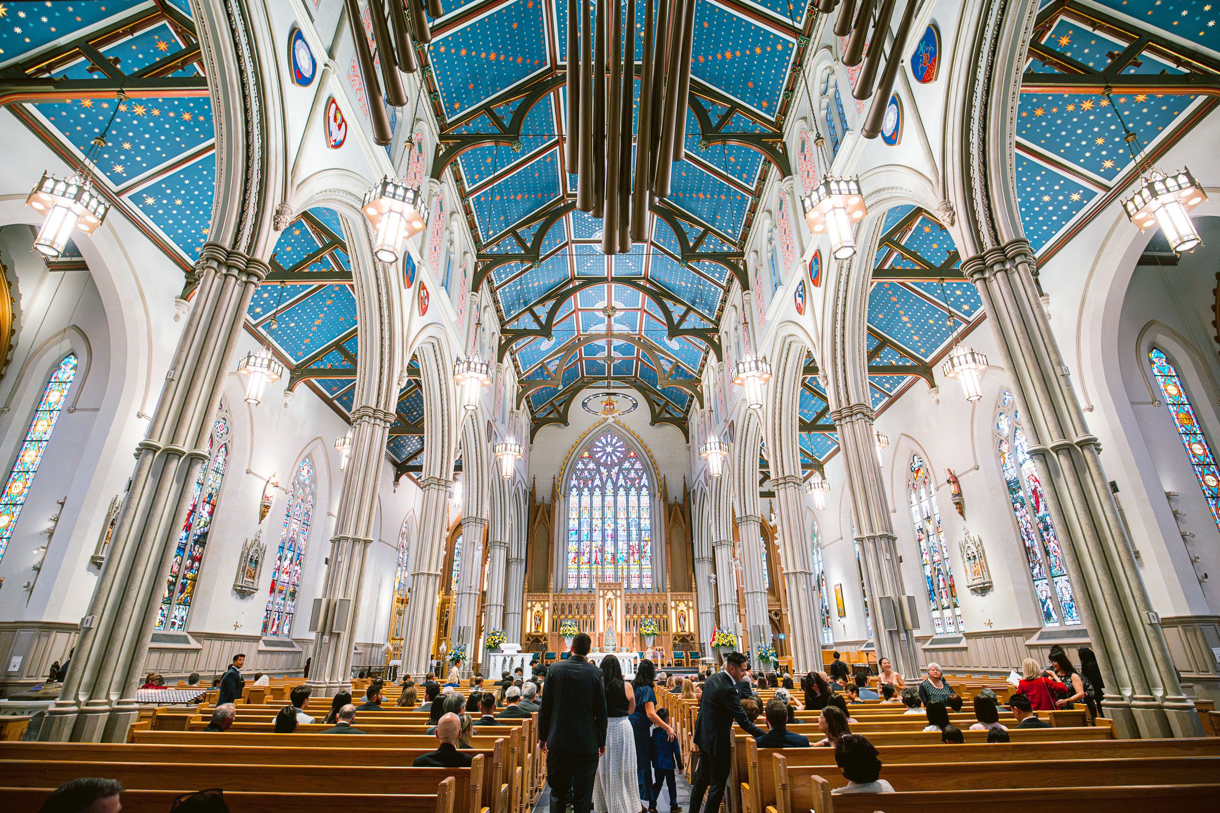Couple walks down the aisle of a grand cathedral with ornate blue ceiling and gothic architecture.