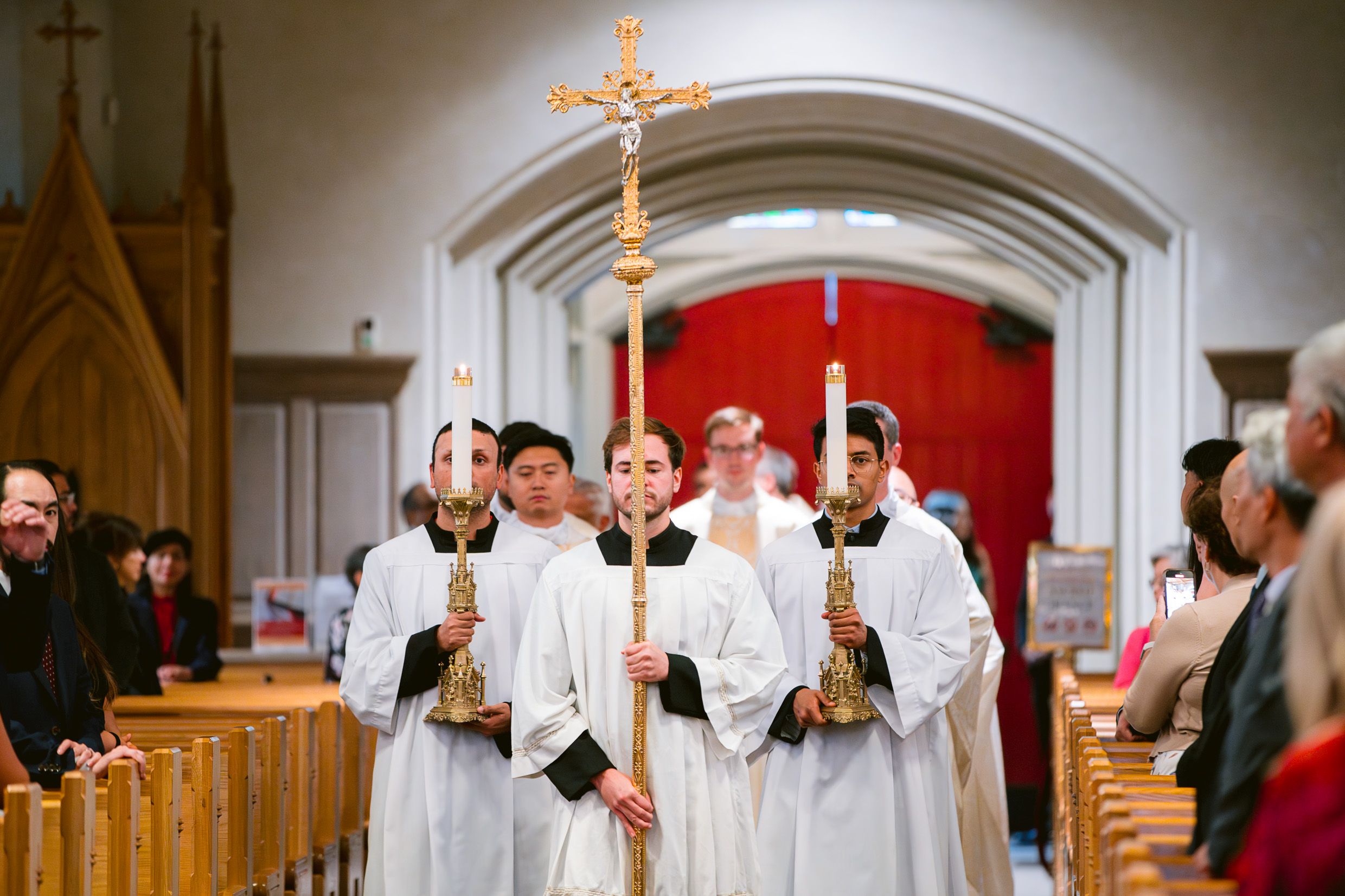 Altar servers in white robes carrying processional crosses and candles during a traditional church ceremony.
