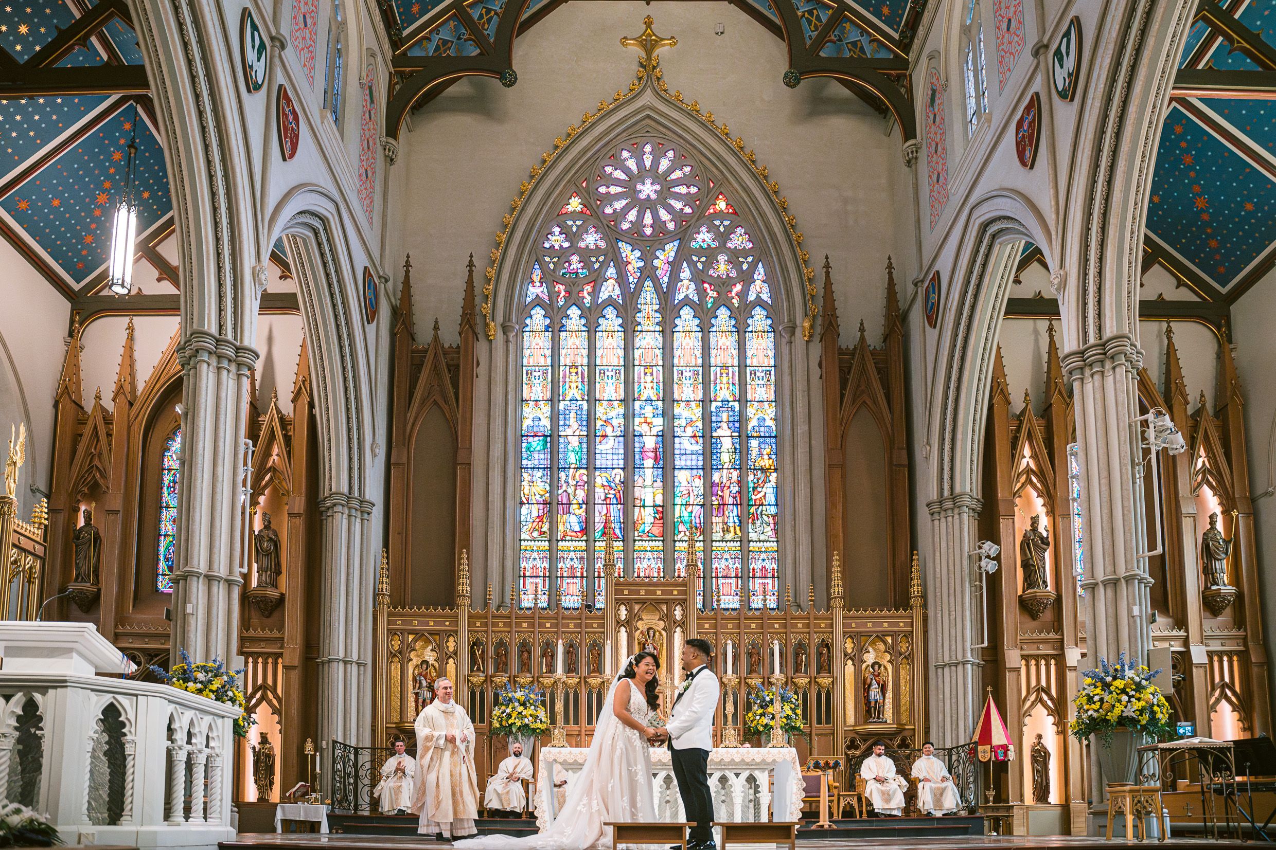 Wedding ceremony taking place inside a grand cathedral with ornate stained glass windows and gothic architecture.
